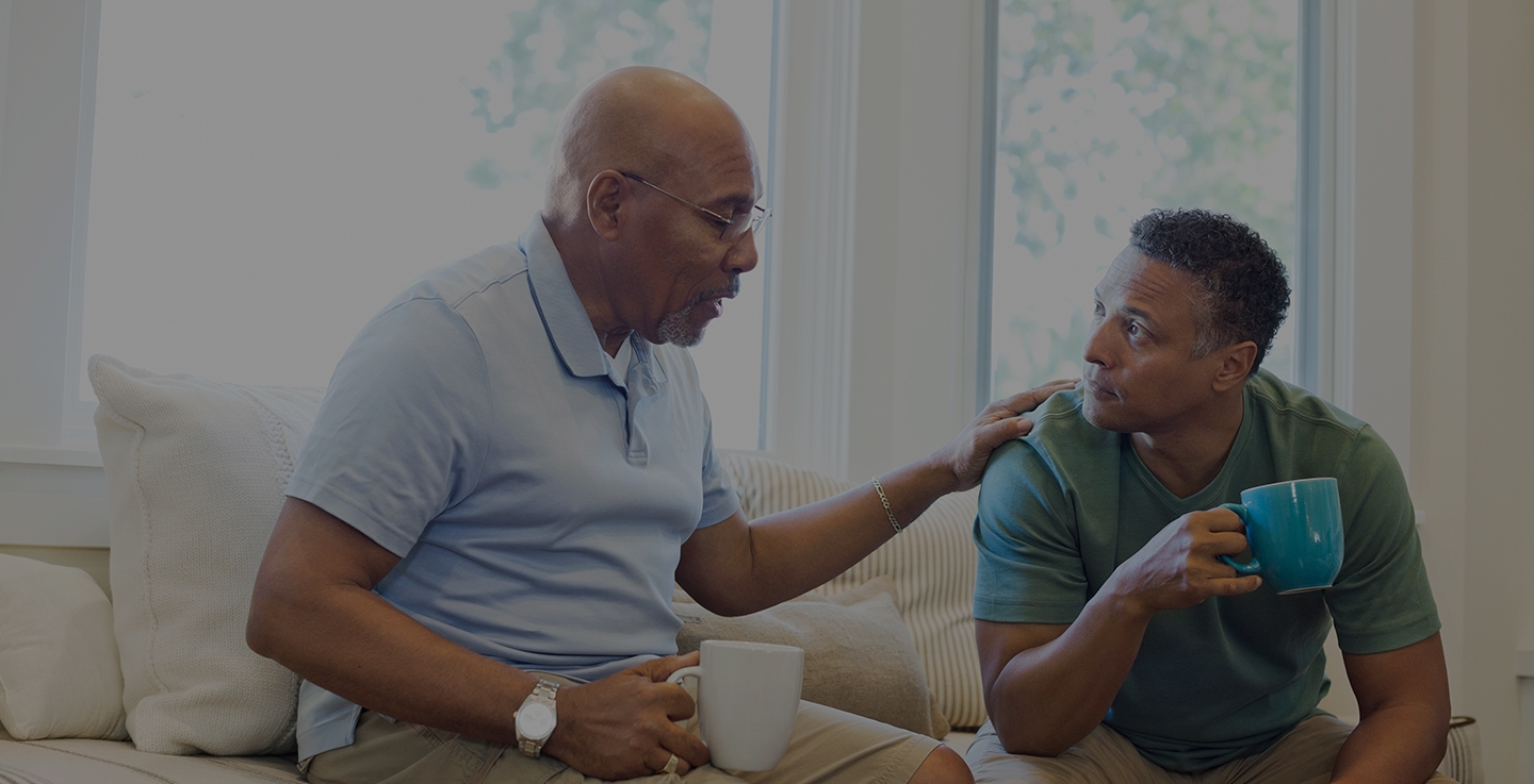 Two men sit in a well-lit living room and discuss cancer care support groups while drinking coffee